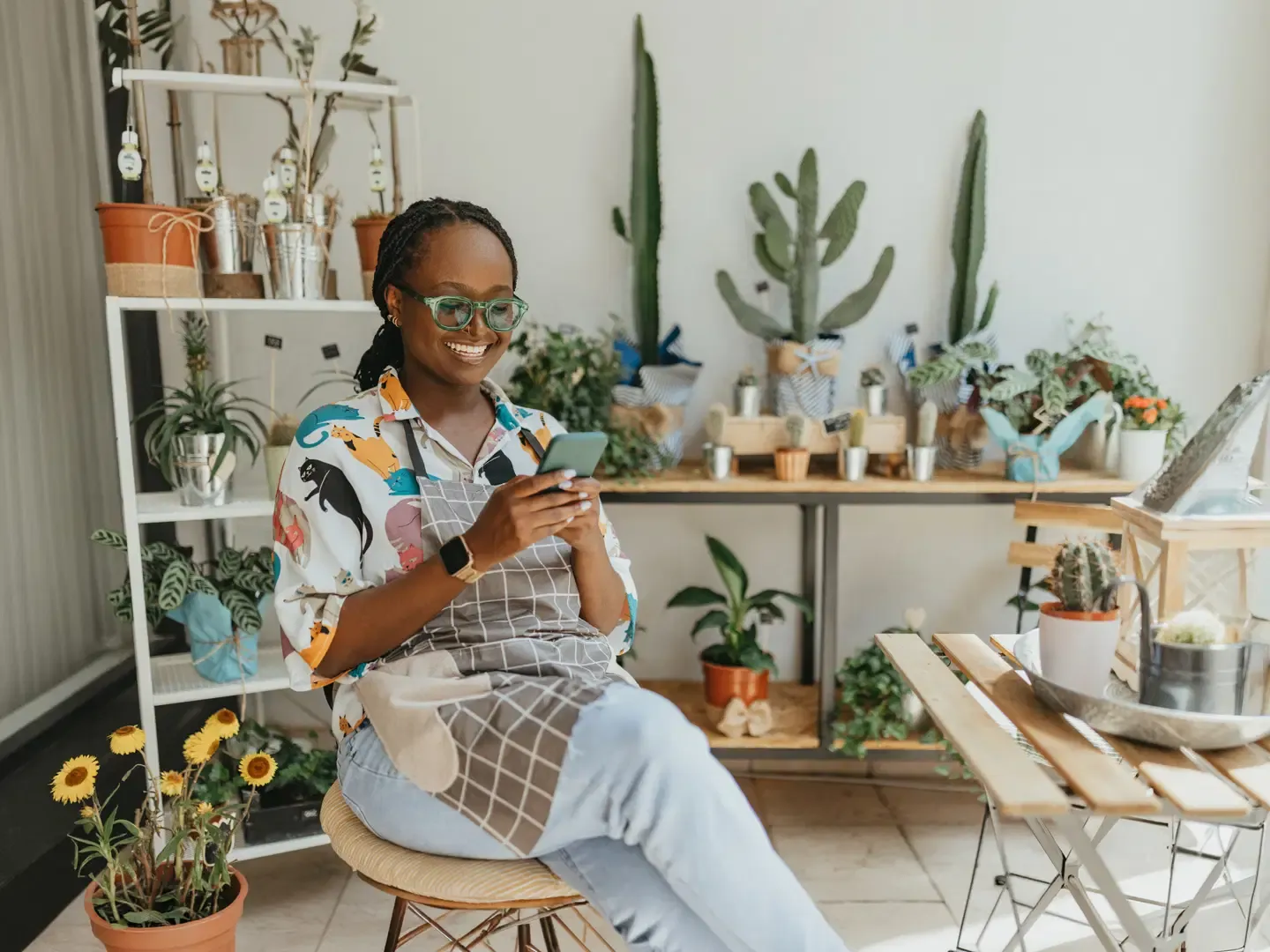 A smiling woman in an apron and glasses sitting in a plant shop while looking at her smartphone.